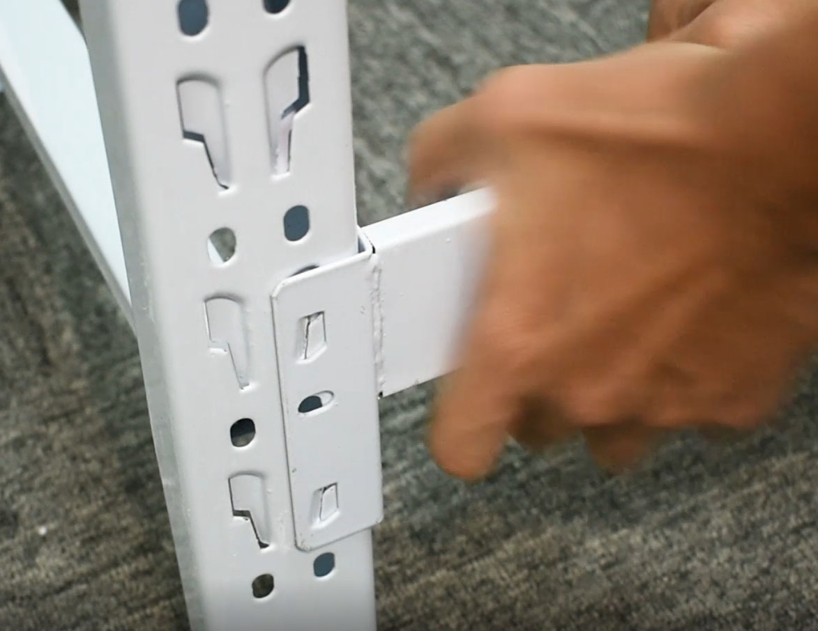 Close-up of a hand pushing the hooked end bracket of a horizontal side frame into the punched slots on the inside face of a white upright