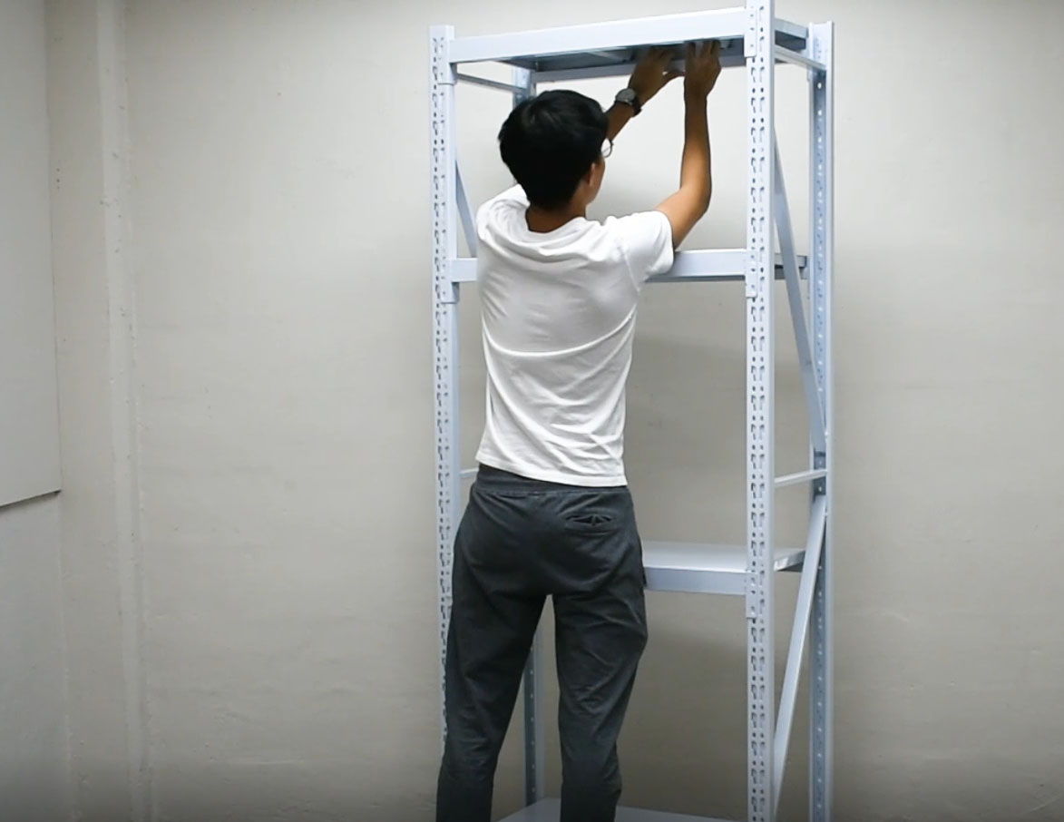 A person laying a metal shelf panel onto the fitted horizontal side frames of a completed white boltless rack, with a lower panel already in place